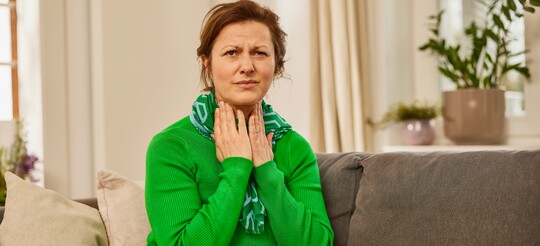 Frau im grünen Pullover sitzt auf dem Sofa und hält sich mit den Händen den Hals, als ob sie Halsschmerzen verspürt. Sie trägt einen Schal und blickt mit einem Ausdruck des Unwohlseins. Die gemütliche Wohnzimmerumgebung mit Pflanzen und Decke vermittelt eine warme, ruhige Atmosphäre für Erholung und Selbstfürsorge.