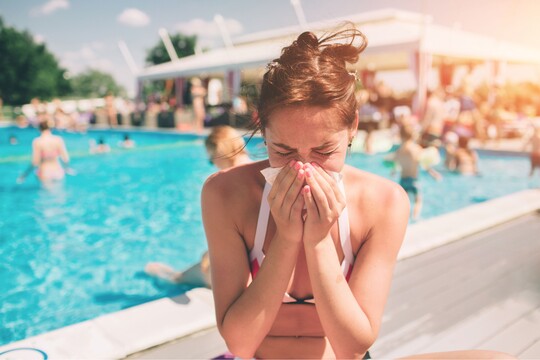 Frau am Pool mit Erkältungssymptomen Junge Frau in Bikini sitzt am Beckenrand eines belebten Pools und schnäuzt sich die Nase, möglicherweise aufgrund einer Erkältung.